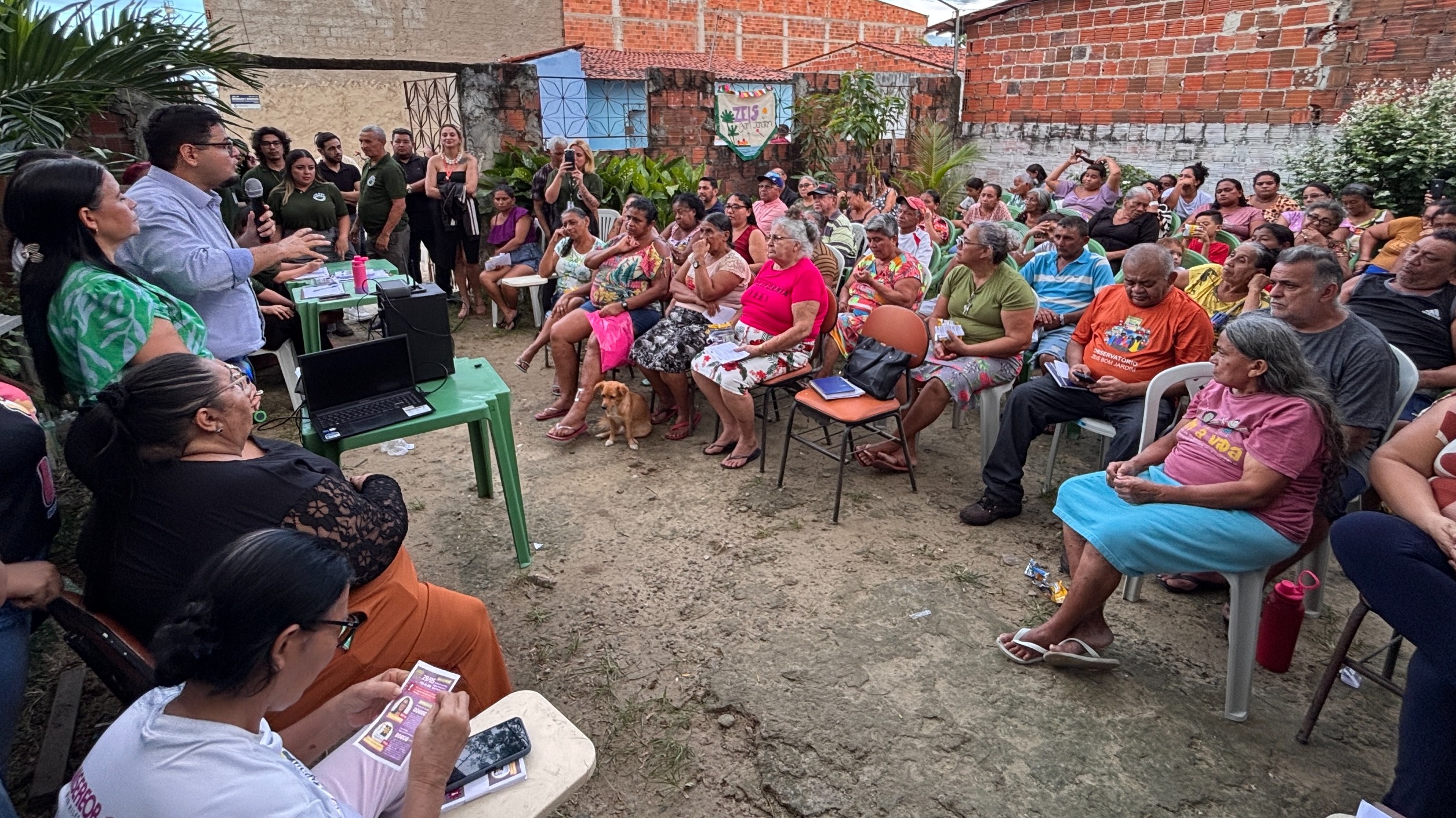 A imagem mostra uma reunião com moradores da comunidade. Eles estão sentados em um circulo ouvindo o secretário da Habitação